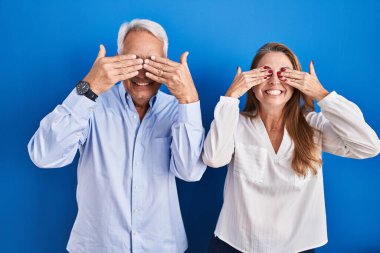 Middle age hispanic couple standing over blue background covering eyes with hands smiling cheerful and funny. blind concept. 