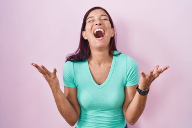 Young hispanic woman standing over pink background celebrating mad and crazy for success with arms raised and closed eyes screaming excited. winner concept 