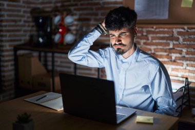Young hispanic man with beard working at the office at night confuse and wonder about question. uncertain with doubt, thinking with hand on head. pensive concept. 