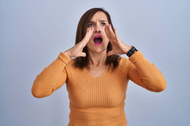 Middle age brunette woman standing wearing orange sweater shouting angry out loud with hands over mouth 