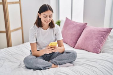 Young hispanic woman using smartphone sitting on bed at bedroom