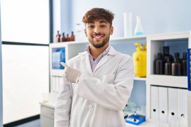 Arab man with beard working at scientist laboratory smiling cheerful pointing with hand and finger up to the side 