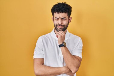 Arab man standing over yellow background with hand on chin thinking about question, pensive expression. smiling and thoughtful face. doubt concept. 