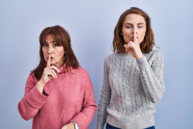 Mother and daughter standing over blue background asking to be quiet with finger on lips. silence and secret concept. 