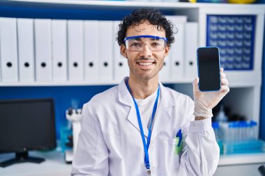 Hispanic man working at scientist laboratory showing smartphone screen looking positive and happy standing and smiling with a confident smile showing teeth 