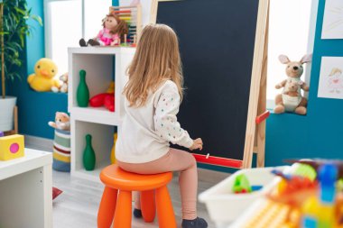 Adorable blonde girl preschool student drawing on blackboard at kindergarten