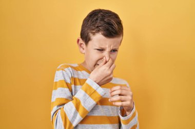 Young caucasian kid standing over yellow background smelling something stinky and disgusting, intolerable smell, holding breath with fingers on nose. bad smell  clipart