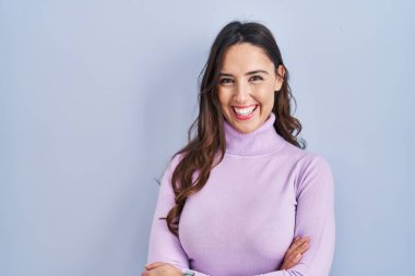 Mavi arka planda duran esmer kadın kollarını kavuşturup kameraya bakarken gülümsüyor. pozitif kişi. 