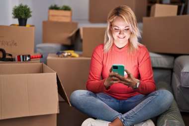 Genç sarışın kadın akıllı telefon kullanıyor. Yeni evinde yerde oturuyor.