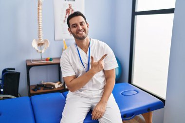 Young hispanic man with beard working at pain recovery clinic cheerful with a smile of face pointing with hand and finger up to the side with happy and natural expression on face 