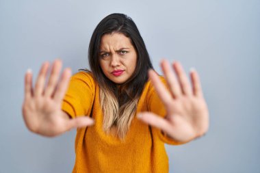 Young hispanic woman standing over isolated background doing stop gesture with hands palms, angry and frustration expression 