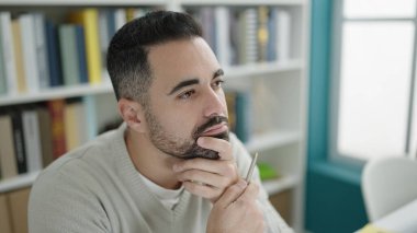 Young hispanic man student sitting on table with doubt expression at library university