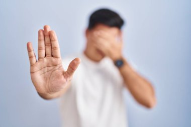 Hispanic man standing over blue background covering eyes with hands and doing stop gesture with sad and fear expression. embarrassed and negative concept. 