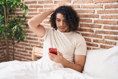 Hispanic man with curly hair using smartphone sitting on the bed stressed and frustrated with hand on head, surprised and angry face 