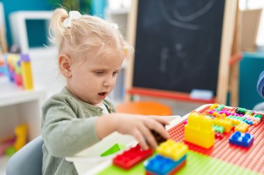 Adorable blonde girl playing with construction blocks sitting on table at kindergarten