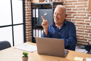 Senior man with grey hair working using computer laptop at the office smiling happy pointing with hand and finger to the side 