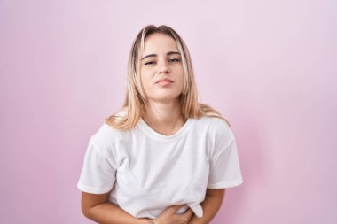 Young blonde woman standing over pink background with hand on stomach because indigestion, painful illness feeling unwell. ache concept. 