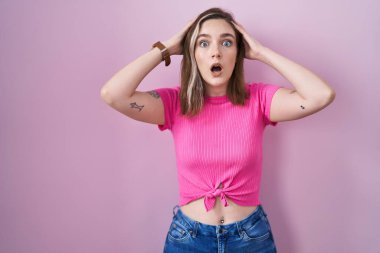 Blonde caucasian woman standing over pink background crazy and scared with hands on head, afraid and surprised of shock with open mouth  clipart