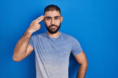 Middle east man with beard standing over blue background shooting and killing oneself pointing hand and fingers to head like gun, suicide gesture. 