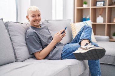 Beyaz bir adam akıllı telefon kullanıyor. Evdeki kanepede oturuyor.