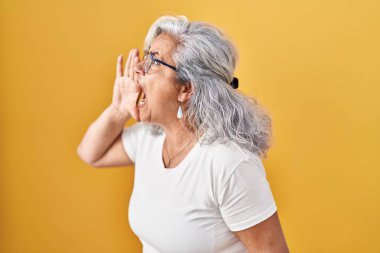 Middle age woman with grey hair standing over yellow background shouting and screaming loud to side with hand on mouth. communication concept. 