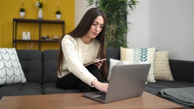 Young beautiful hispanic woman writing on notebook using laptop at home