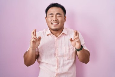 Chinese young man standing over pink background gesturing finger crossed smiling with hope and eyes closed. luck and superstitious concept. 