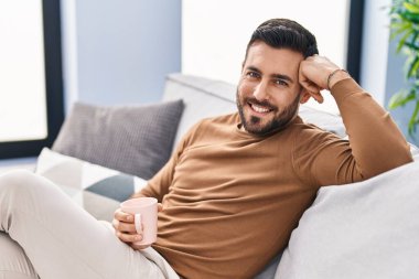 Genç İspanyol adam evdeki kanepede kahve içiyor.