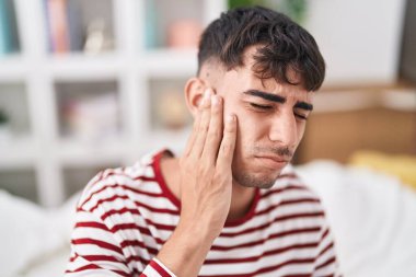 Young hispanic man suffering for ear pain sitting on bed at bedroom