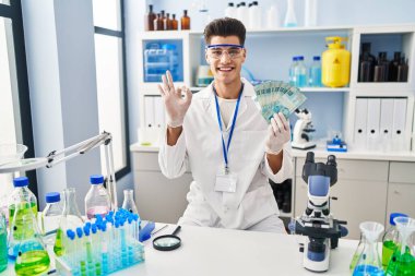 Young hispanic man working at scientist laboratory holding brazilian reals doing ok sign with fingers, smiling friendly gesturing excellent symbol 
