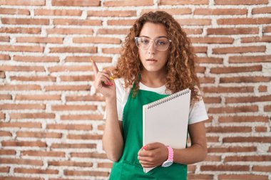 Young caucasian woman holding art notebook pointing with hand finger to the side showing advertisement, serious and calm face 