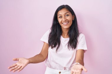 Young hispanic woman standing over pink background smiling cheerful with open arms as friendly welcome, positive and confident greetings 