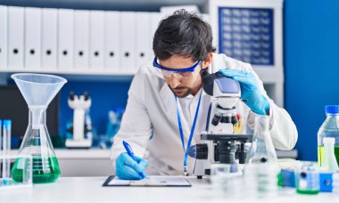 Young hispanic man scientist using microscope write on clipboard at laboratory