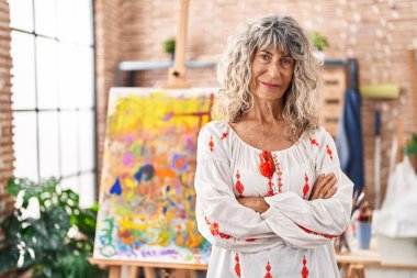Middle age woman artist standing with arms crossed gesture at art studio