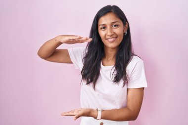 Young hispanic woman standing over pink background gesturing with hands showing big and large size sign, measure symbol. smiling looking at the camera. measuring concept. 