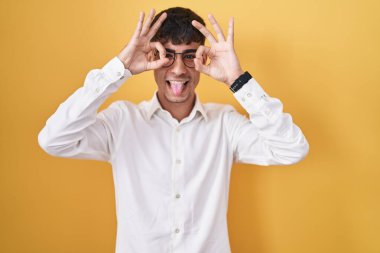 Sarı arka planda dikilen İspanyol genç adam dürbün gibi jest yapıyor. Dürbün dilini çıkarıyor, gözler parmaklarından bakıyor. çılgın ifade. 