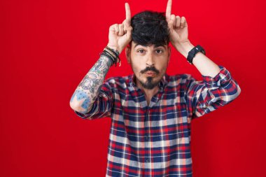 Young hispanic man with beard standing over red background doing funny gesture with finger over head as bull horns 