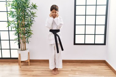 Young hispanic girl wearing karate kimono and black belt covering ears with fingers with annoyed expression for the noise of loud music. deaf concept. 