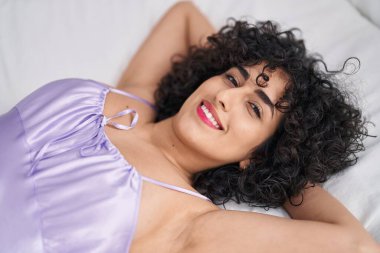 Orta Doğulu genç kadın, elleri başının üstünde, yatak odasında uzanıyor.