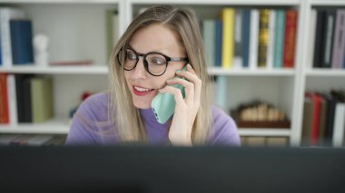 Young blonde woman student using computer talking on smartphone at library university