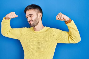 Hispanic man standing over blue background showing arms muscles smiling proud. fitness concept. 