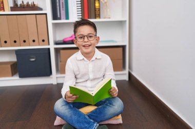 Adorable hispanic boy student smiling confident reading book at library school