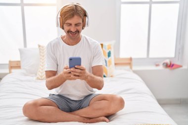 Young blond man listening to music sitting on bed at bedroom