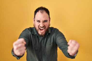 Plus size hispanic man with beard standing over yellow background angry and mad raising fists frustrated and furious while shouting with anger. rage and aggressive concept. 