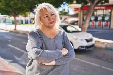 Middle age blonde woman standing relaxed with arms crossed gesture at street