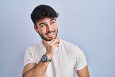 Hispanic man with beard standing over white background with hand on chin thinking about question, pensive expression. smiling and thoughtful face. doubt concept. 