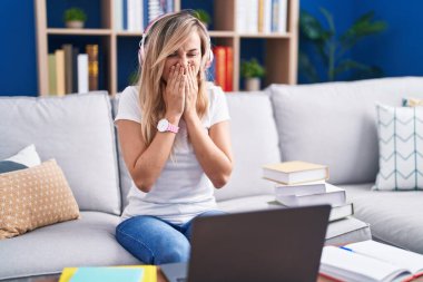 Young blonde woman studying using computer laptop at home laughing and embarrassed giggle covering mouth with hands, gossip and scandal concept 