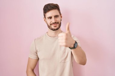 Hispanic man with beard standing over pink background looking proud, smiling doing thumbs up gesture to the side 