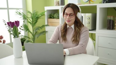 Young beautiful hispanic woman using laptop and headphones sitting on table at home