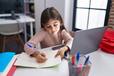 Adorable hispanic girl student using laptop writing on notebook at classroom